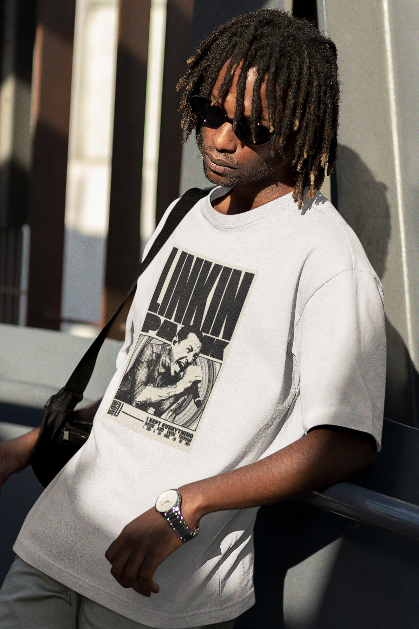 Person wearing a white t-shirt with graphic design, sunglasses, and a watch, leaning against a wall.